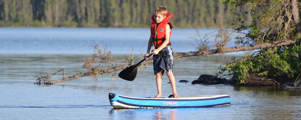A boy in a life jacket paddle boarding on calm water, showcasing that paddle boarding anyone can do it.