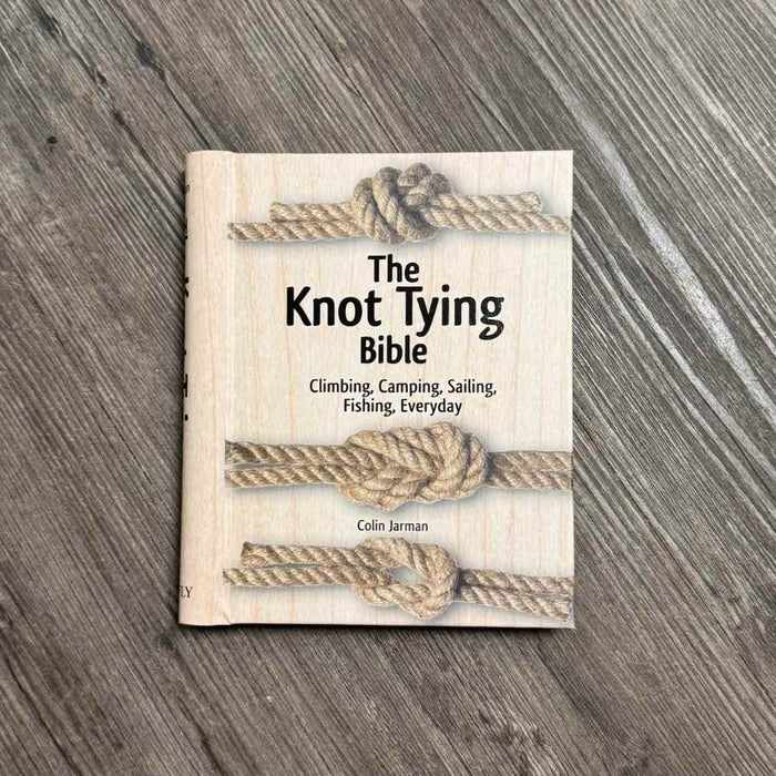 The Knot Tying Bible book by Colin Jarman, featuring a light wood-grain cover with three illustrated rope knots, placed on a gray wood-textured surface.