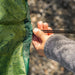 A close-up of a person's hand adjusting the drawstring closure of a green camping tablecloth with outdoor gear illustrations, set against a blurred natural background.