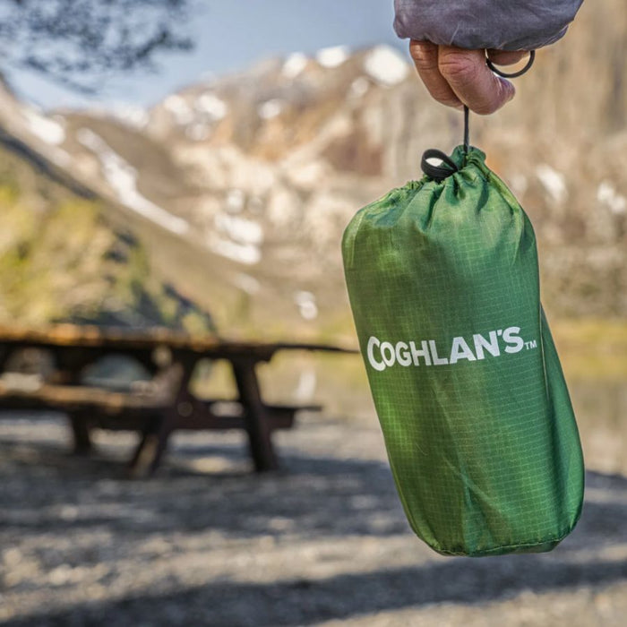 A green drawstring bag with the brand name 'COGHLAN'S' printed in white, held by a hand against a blurred mountain and picnic table background.