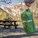 A green drawstring bag with the brand name 'COGHLAN'S' printed in white, held by a hand against a blurred mountain and picnic table background.