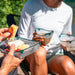 A lifestyle shot of a group outdoors sharing a picnic with clear glass bowls featuring a green striped pattern and a white base, holding food like crackers, cheese, and berries.