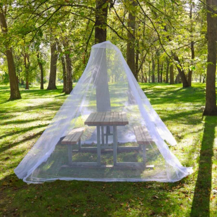 A white mosquito net tent draped over a wooden picnic table in a sunlit park with green grass and trees.