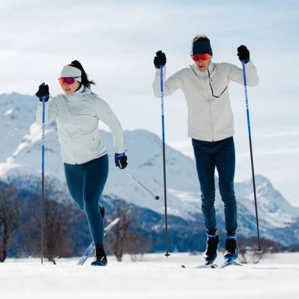 Two people wearing navy blue pants and white jackets skiing in a snowy, mountainous landscape.