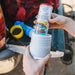 A person's hands holding a white plastic bottle container and inserting a cylindrical canister labeled 'Counter Assault' into it, with a blurred outdoor background.
