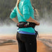 A woman wearing a turquoise long-sleeve athletic shirt and black leggings is shown from behind outdoors, wearing a black waist pack with red and white accents. The background shows a misty natural landscape.