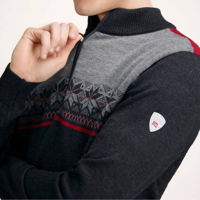 A close-up of a man wearing a dark gray and gray heather knit sweater with a patterned yoke, red accents, and a small shield-shaped logo patch on the sleeve.