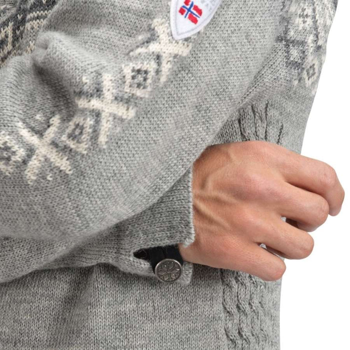 Close-up of a person wearing a gray knit sweater with cream Nordic-style patterns, a white shield patch with the Norwegian flag, and a black button closure.