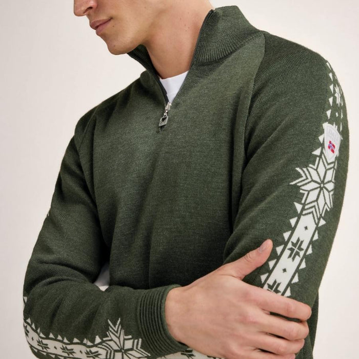A man wearing an olive green knit quarter-zip sweater with white Nordic snowflake patterns on the sleeves, shown from the chest down against a light neutral background.