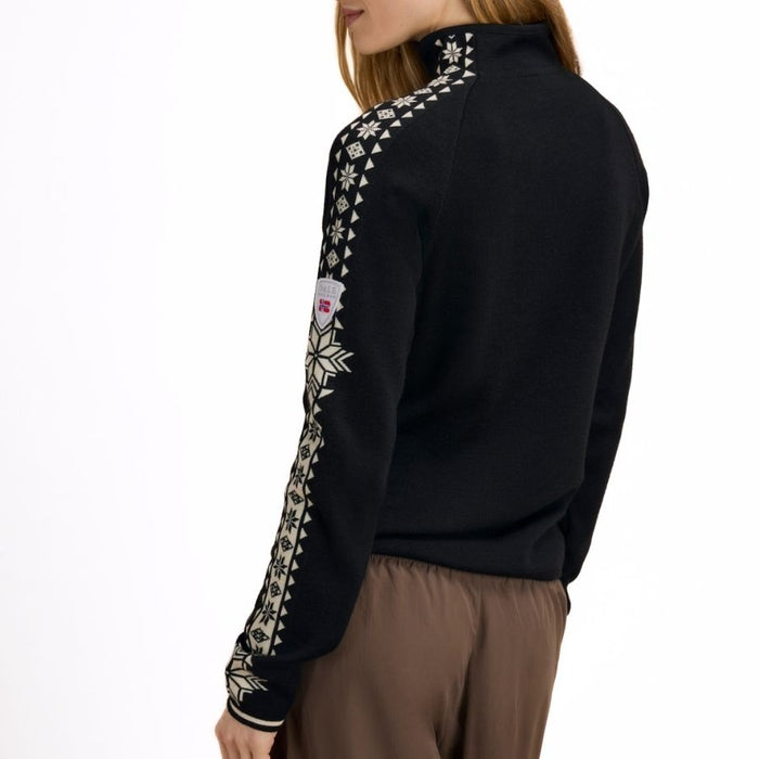 A woman shown from the side wearing a black mock-neck knit sweater with intricate white geometric and snowflake patterns down the sleeves, paired with brown trousers, against a plain white background.