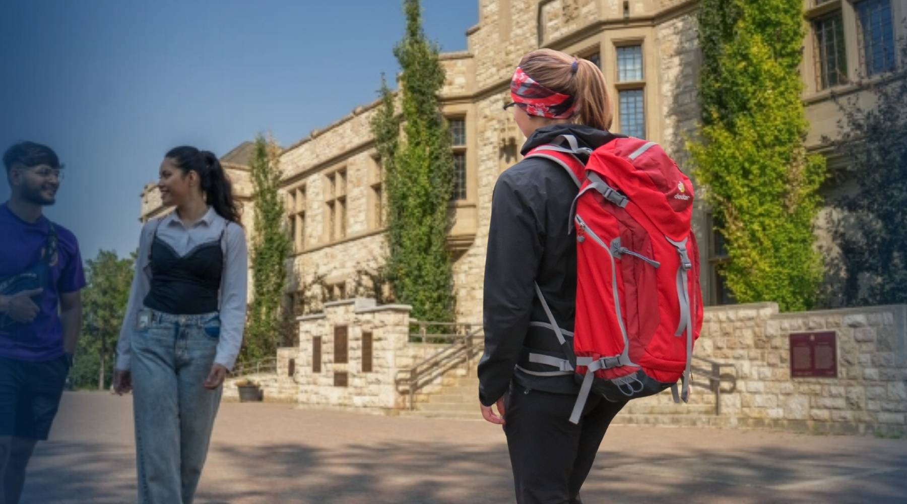 A woman wearing a red hiking backpack walks outdoors in front of a stone building, with two other people visible in the background.