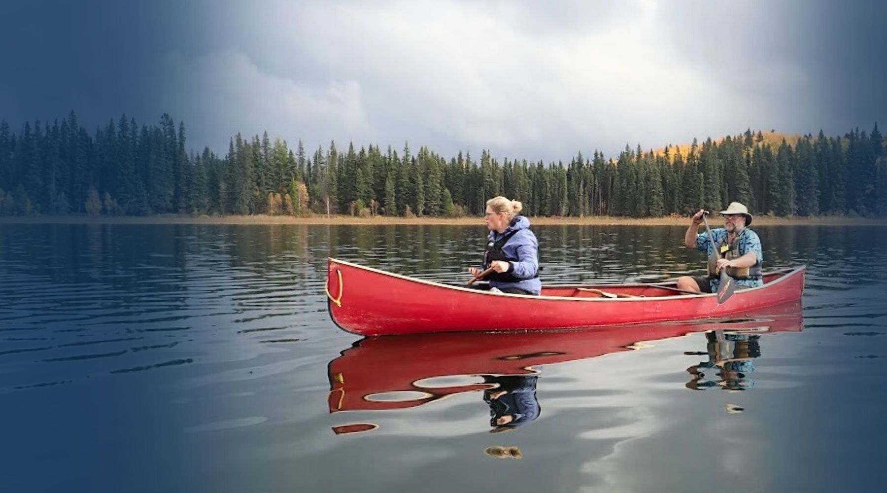 A red canoe floats on calm lake water with a forest shoreline under a cloudy sky. Two people are seated in the canoe, one facing forward and one looking out to the side.