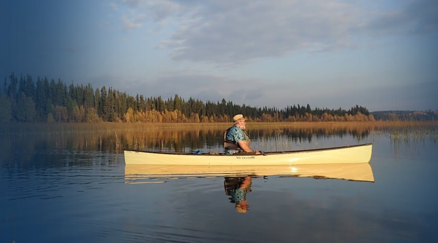 A person sits in a light-colored canoe on a calm lake, with a forest shoreline and blue sky reflected in the water.