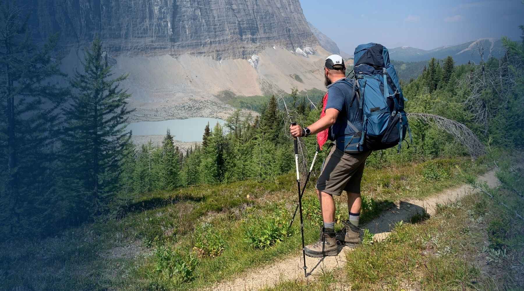 A hiker stands on a trail in a mountain landscape, wearing a large blue hiking backpack and using trekking poles. Trees, grass, a lake, and a rocky mountain are visible in the background.