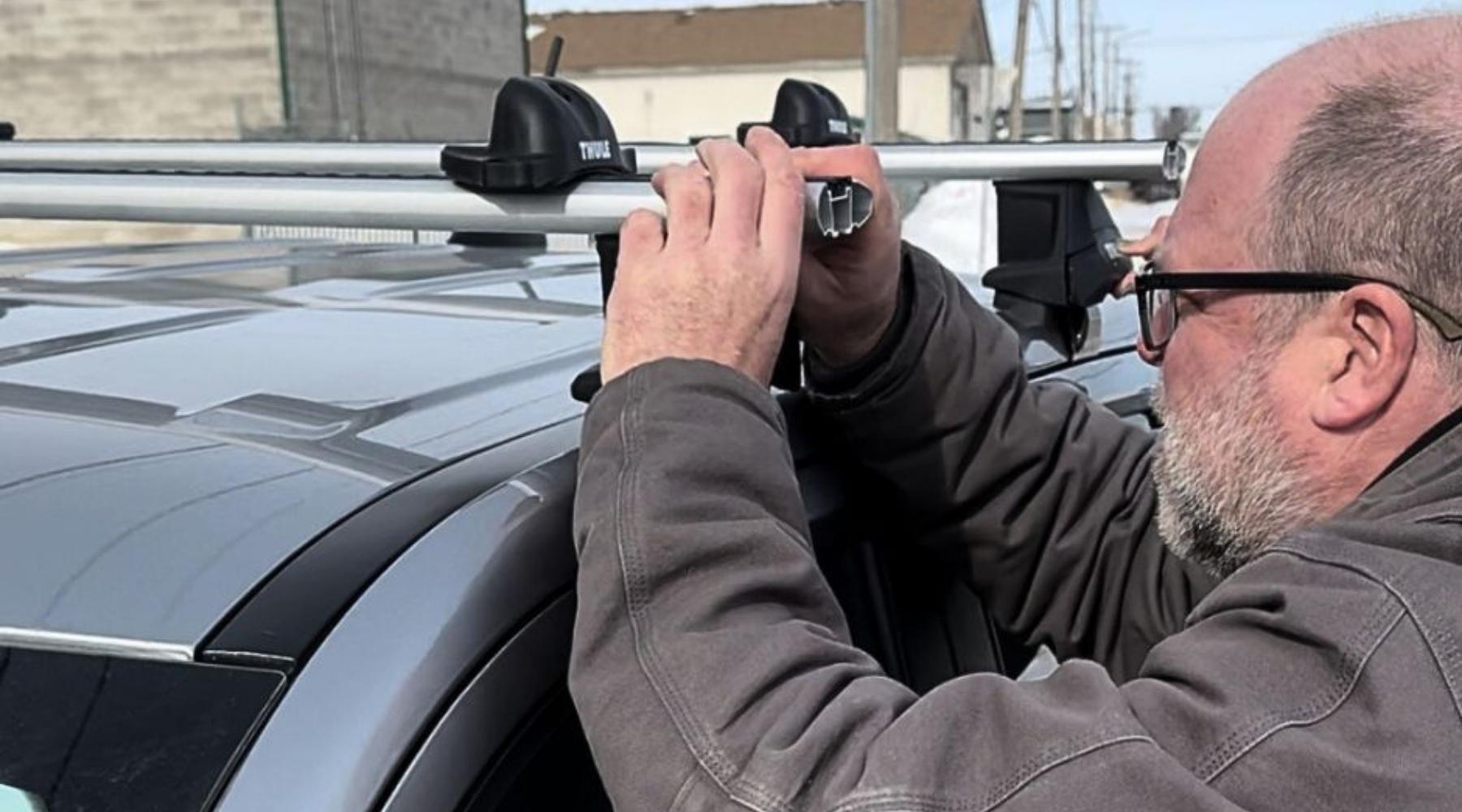 A man wearing glasses and a brown jacket is installing a roof rack onto the roof of a gray vehicle outdoors.