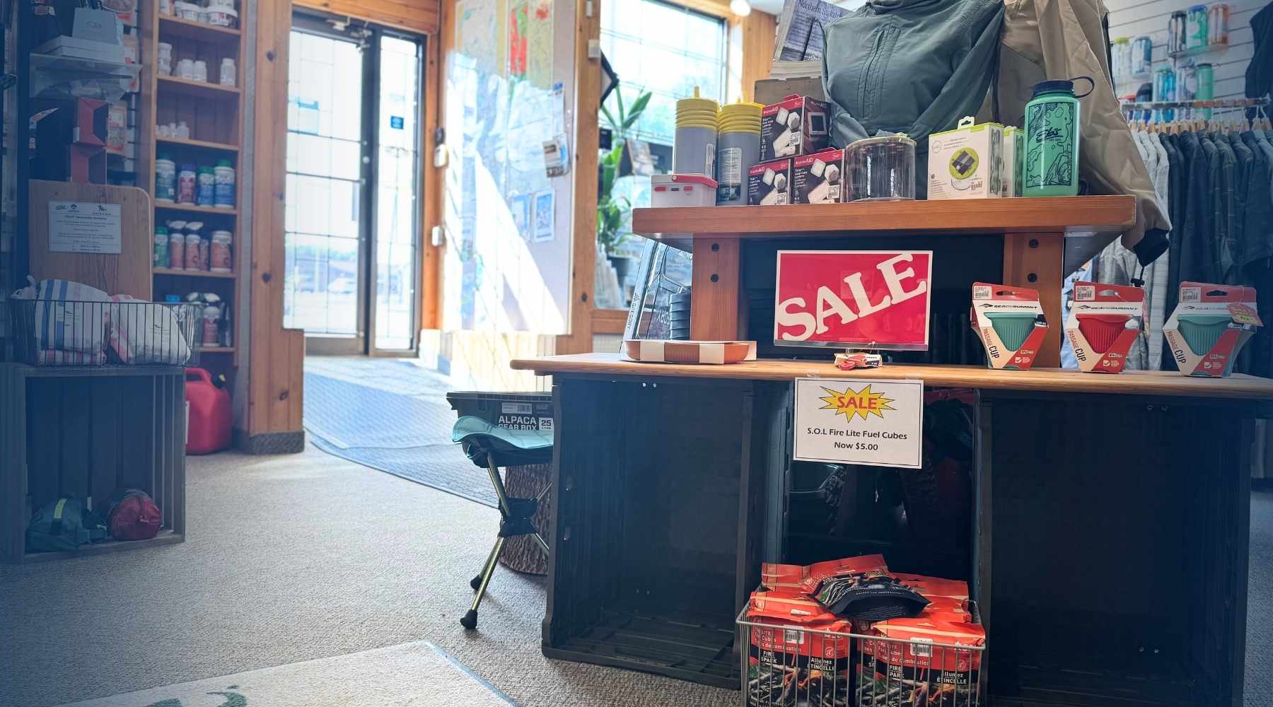 A retail store interior with a wooden display table holding a red SALE sign, packaged goods, and merchandise. Shelves, glass doors, and clothing racks are visible in the background.