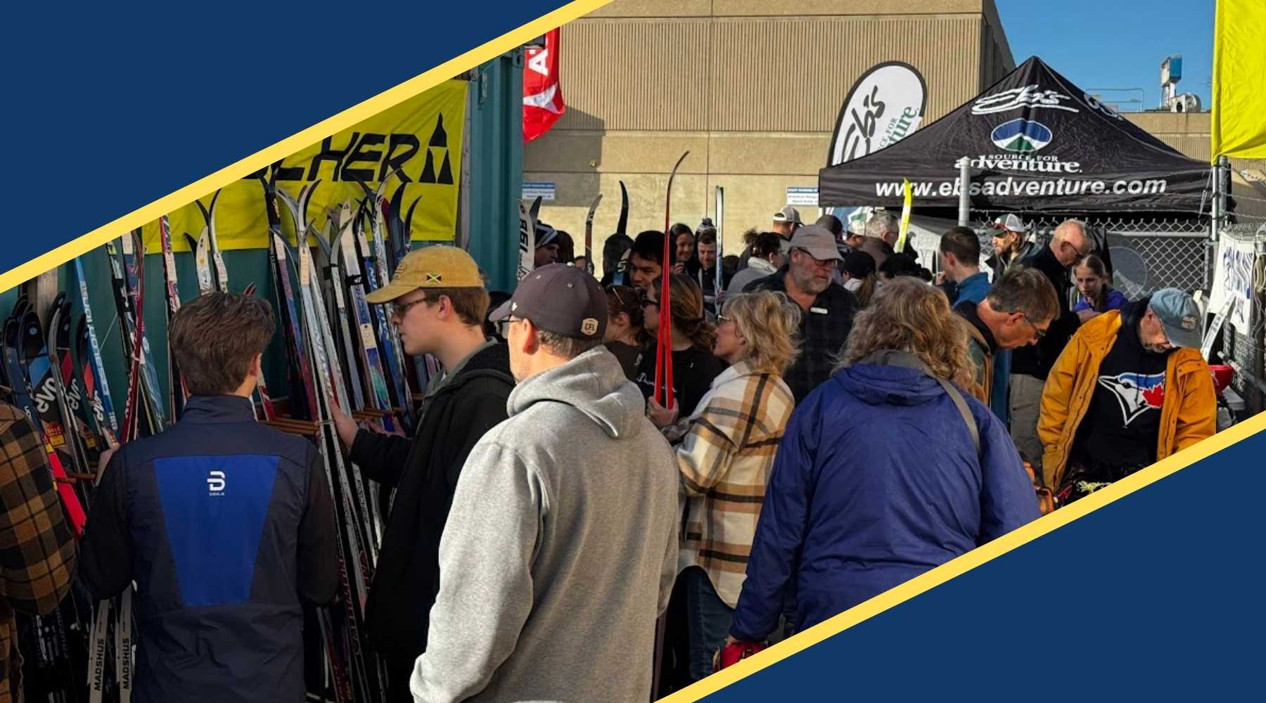 A crowded outdoor ski event with people browsing racks of skis and standing near branded tents, including a black tent with the text 'Ebs Adventure' and 'www.ebsadventure.