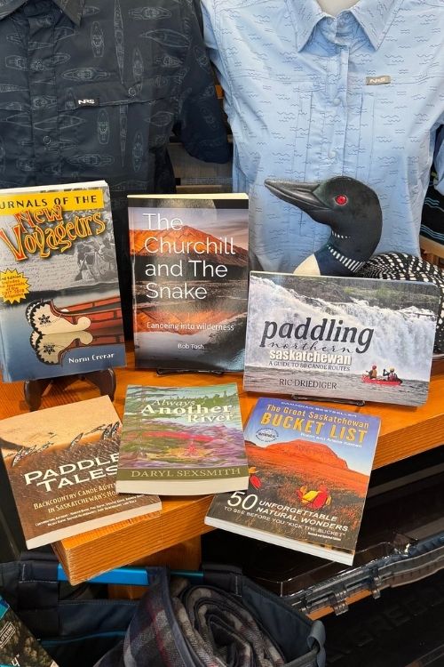A retail display table featuring a collection of books, including titles like 'The Churchill and the Snake', 'paddling with the Southern Lake', and 'Paddle Tales', alongside a loon figurine and shirts on mannequins in the background.