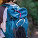 A teal blue hiking backpack worn by a person outdoors, with a pink strap and a white carabiner attached, and a pair of black hiking boots hanging from the side.