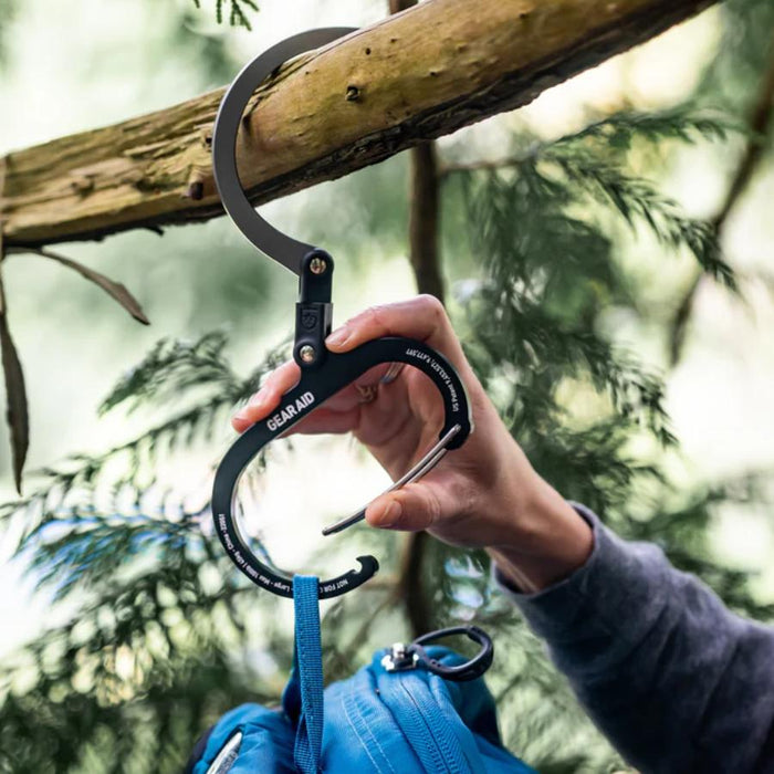 A hand holding a black Gearaid quick-release climbing carabiner with a blue climbing rope attached, set against a blurred forest background with a tree branch overhead.