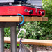 A red portable camping stove sits on a wooden picnic table outdoors, with a blue and black carabiner clip holding a gray bag below the table.