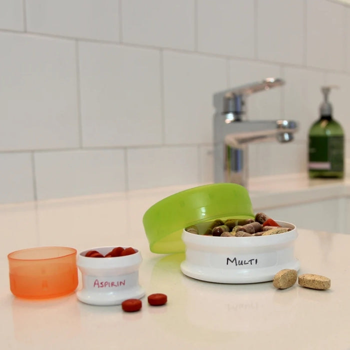 A set of small white pill organizer containers with colorful lids, including one labeled 'Aspirin' with red tablets, one labeled 'Multi' with mixed tablets, and an empty orange container, arranged on a bathroom counter with a sink and tiled wall in the background.