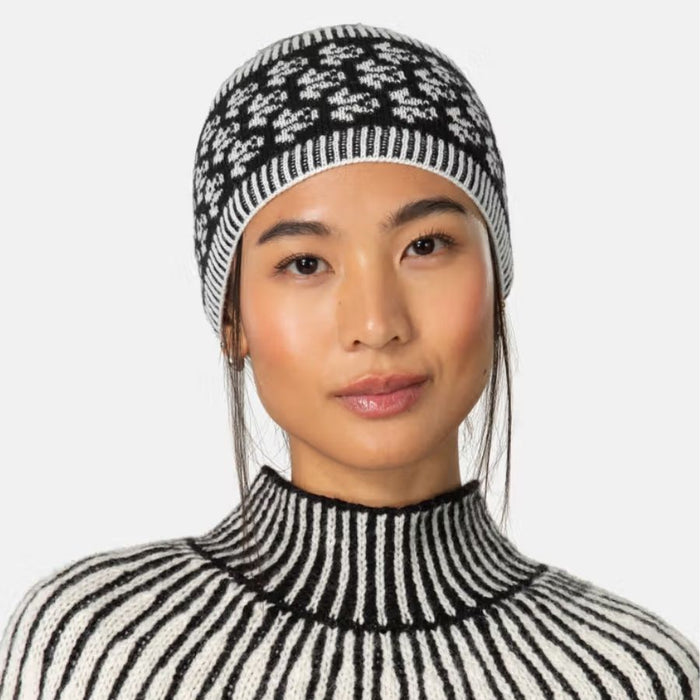 A woman wearing a black and white knit beanie with a snowflake pattern and a matching black and white striped turtleneck sweater, shown against a light gray background.