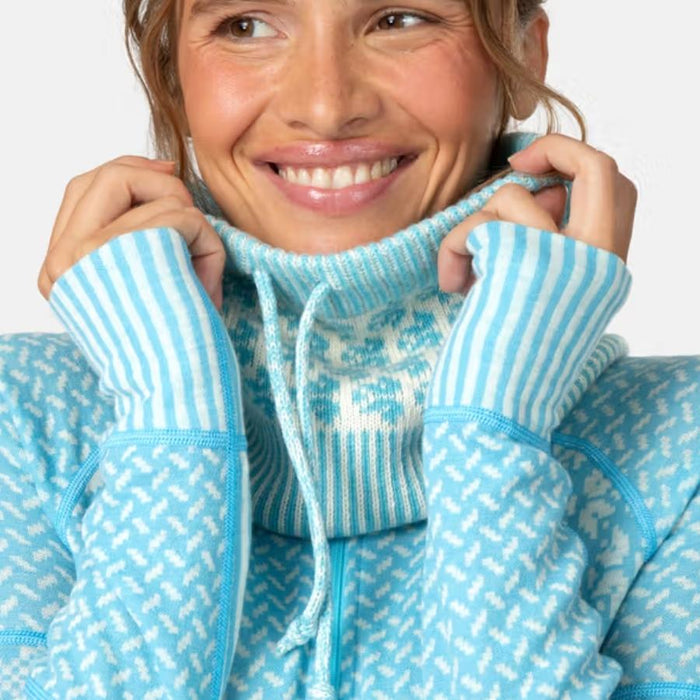 A close-up of a smiling woman wearing a light blue and white patterned fleece hoodie with a high ribbed turtleneck collar, holding the collar up with both hands.