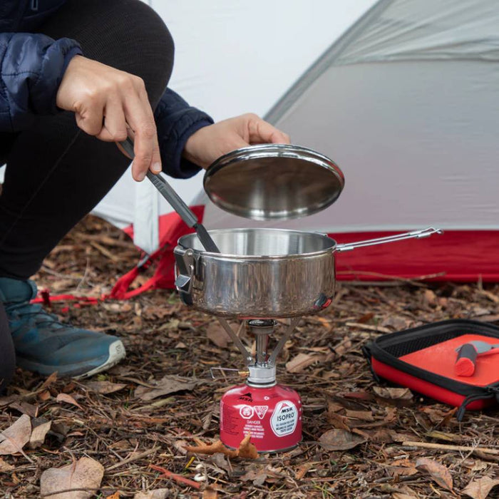 A stainless steel camping pot on a small portable gas stove, set on a red fuel canister outdoors in front of a tent. A person is crouching and holding the pot lid, with a red and black carrying case on the ground nearby.