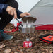 A stainless steel camping pot on a small portable gas stove, set on a red fuel canister outdoors in front of a tent. A person is crouching and holding the pot lid, with a red and black carrying case on the ground nearby.