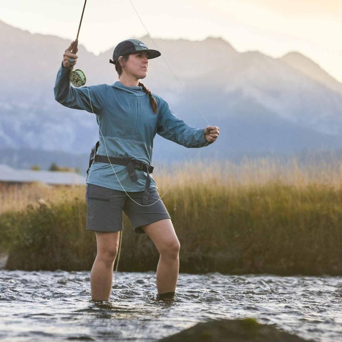 A woman fly fishing in a river, wearing a teal long-sleeve performance shell, dark gray shorts, and a black cap, with mountains in the background.