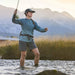 A woman fly fishing in a river, wearing a teal long-sleeve performance shell, dark gray shorts, and a black cap, with mountains in the background.