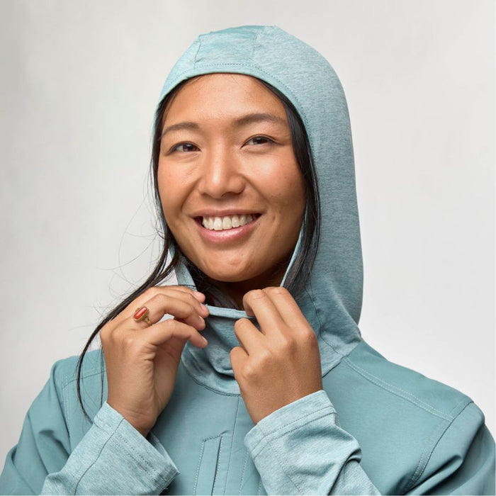 A smiling woman wearing a light blue hooded jacket, adjusting the hood with both hands against a plain light gray background.