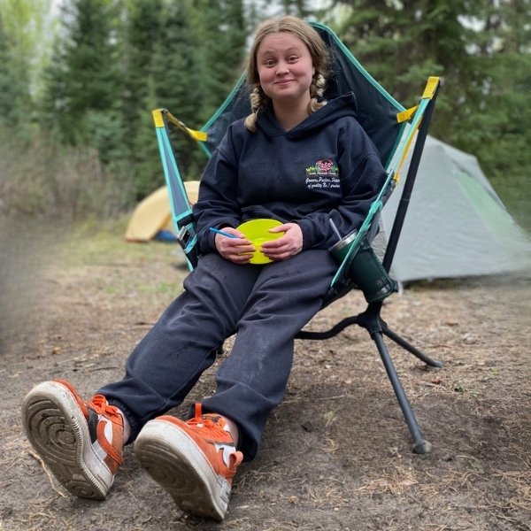 A young girl sits in a teal and yellow camping chair outdoors on a campsite, wearing a navy hoodie with a 'Proud of Our Camp' logo, navy pants, and orange sneakers. A tent is visible in the background among pine trees.