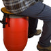 A person wearing a plaid shirt and dark pants sits on a red plastic barrel with a yellow seat pad and black harness straps, isolated on a white background.