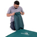 A man wearing a gray t-shirt and glasses kneels on a white studio background, holding a dark green Sea to Summit bag next to a matching dark green sleeping pad with the Sea to Summit logo.