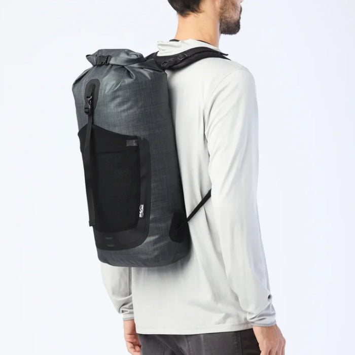 A man wearing a light gray long-sleeve shirt and dark pants, shown from the back and side, wearing a dark gray waterproof backpack with a black front pocket and black straps against a white studio background.