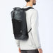 A man wearing a light gray long-sleeve shirt and dark pants, shown from the back and side, wearing a dark gray waterproof backpack with a black front pocket and black straps against a white studio background.