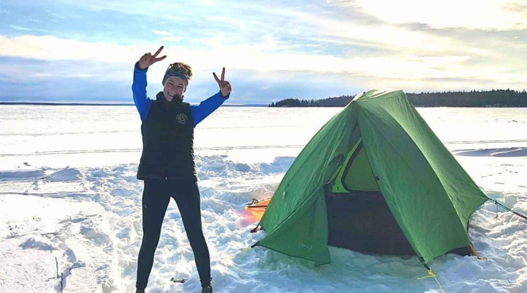 Woman giving the peace sign standing on a snowy lake beside a green tent.