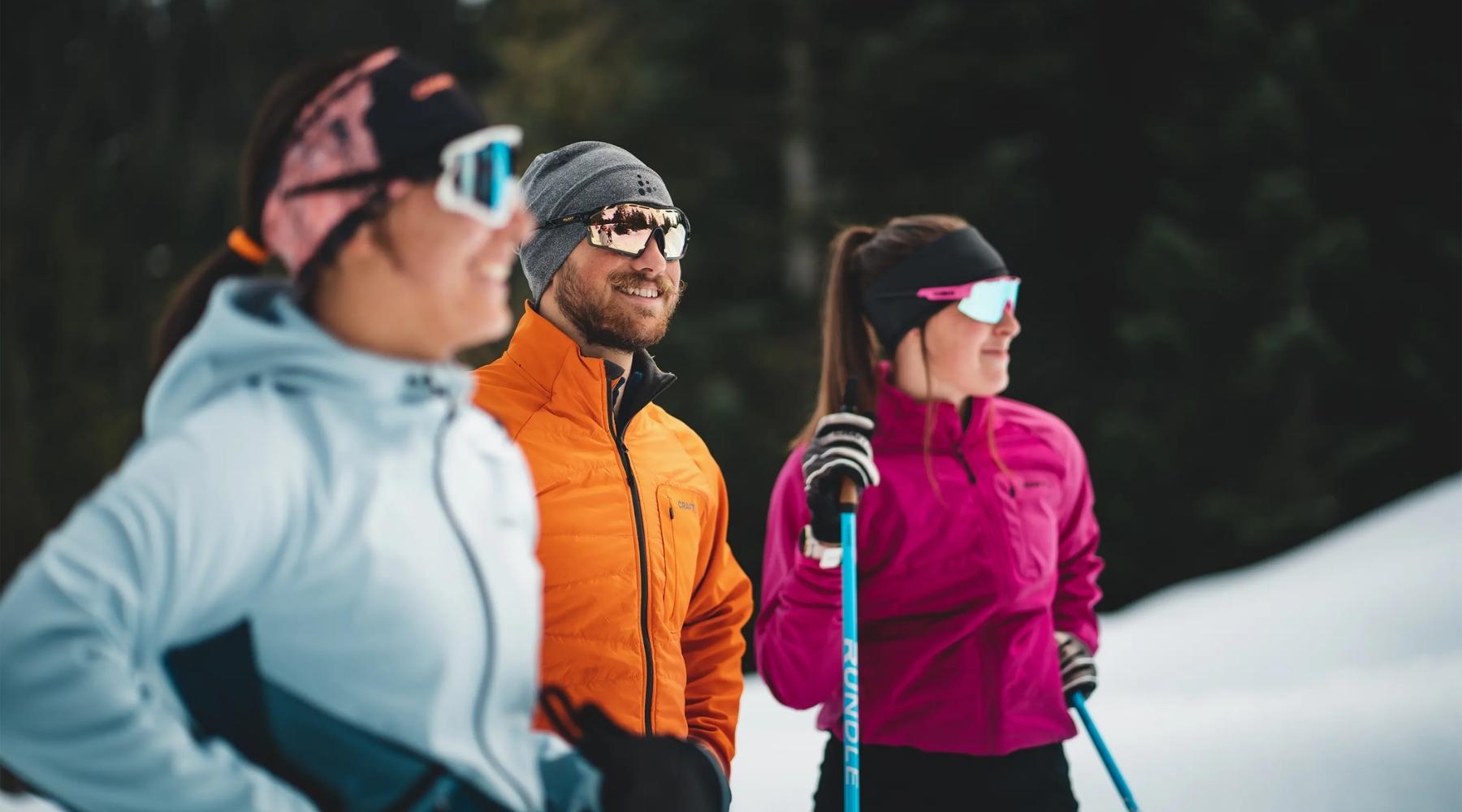 Three people in colourful jackets and ski poles against a snowy forested background.
