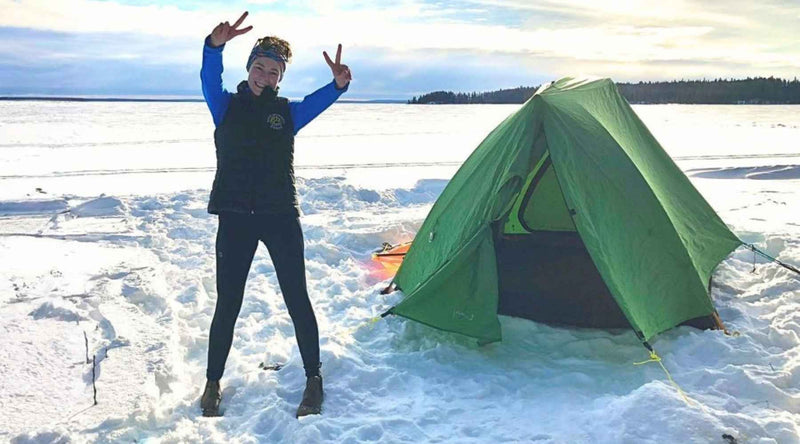 Woman standing on a frozen lake beside a green tent, giving the peace sign with both hands.