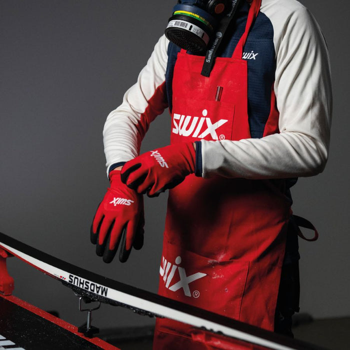 Person wearing a red Swix apron and gloves, standing next to a ski with a gray background.