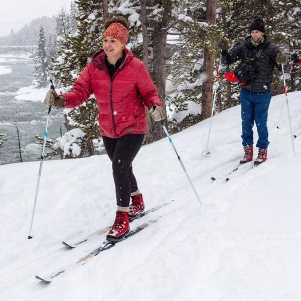 Two people skiing through a snowy wooded landscape with a river in the background, one wearing a red jacket.