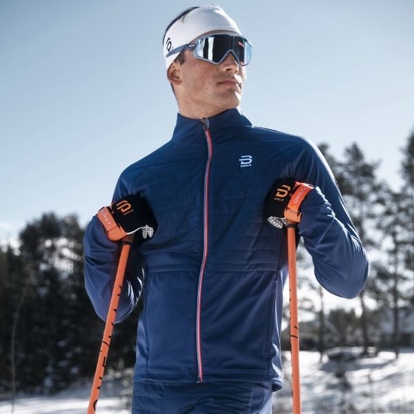 Man wearing a navy jacket and holding orange ski poles against a snowy wooded landscape.