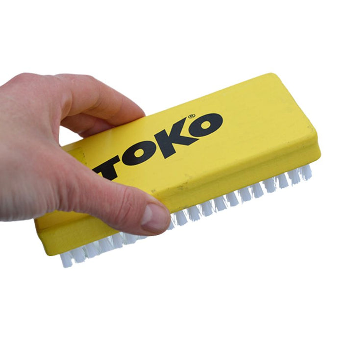 A hand holding a yellow TOKO plastic cleaning brush with white bristles against a plain white background.