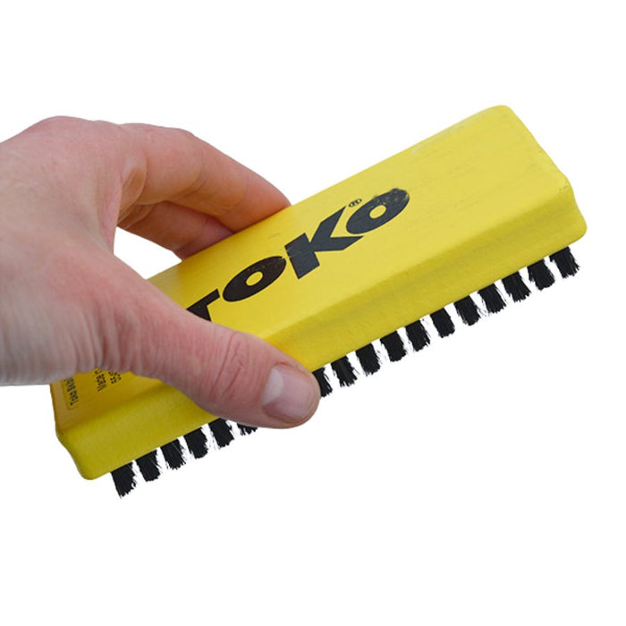 A hand holding a yellow TOKO horsehair brush with black bristles against a plain white background.