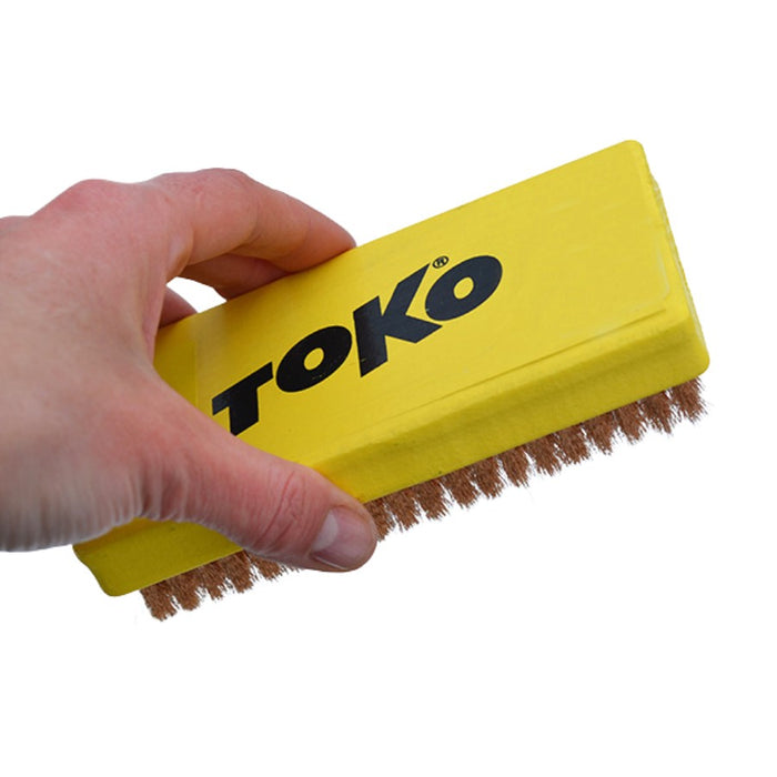 A hand holding a yellow rectangular cleaning brush with bronze bristles and the brand name TOKO printed in black on the side, against a plain white background.