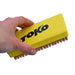 A hand holding a yellow rectangular cleaning brush with bronze bristles and the brand name TOKO printed in black on the side, against a plain white background.