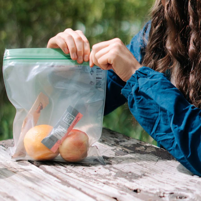 A person wearing a blue jacket is shown from the side, opening a clear plastic zip-top bag with a green seal on a rustic wooden table outdoors. Inside the bag are an orange and a packaged snack stick.